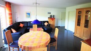 Un salon avec un canapé noir, une table ronde recouverte d’une nappe à carreaux et une lampe suspendue bleue.
