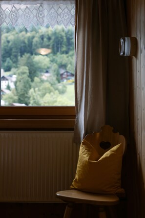 Una ventana con vistas a la vegetación, un radiador, una silla con cojín y una cortina.