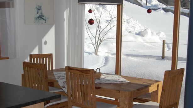 Uma mesa de jantar de madeira com quatro cadeiras, uma toalha de mesa florida e vista para uma paisagem coberta de neve através de uma janela.