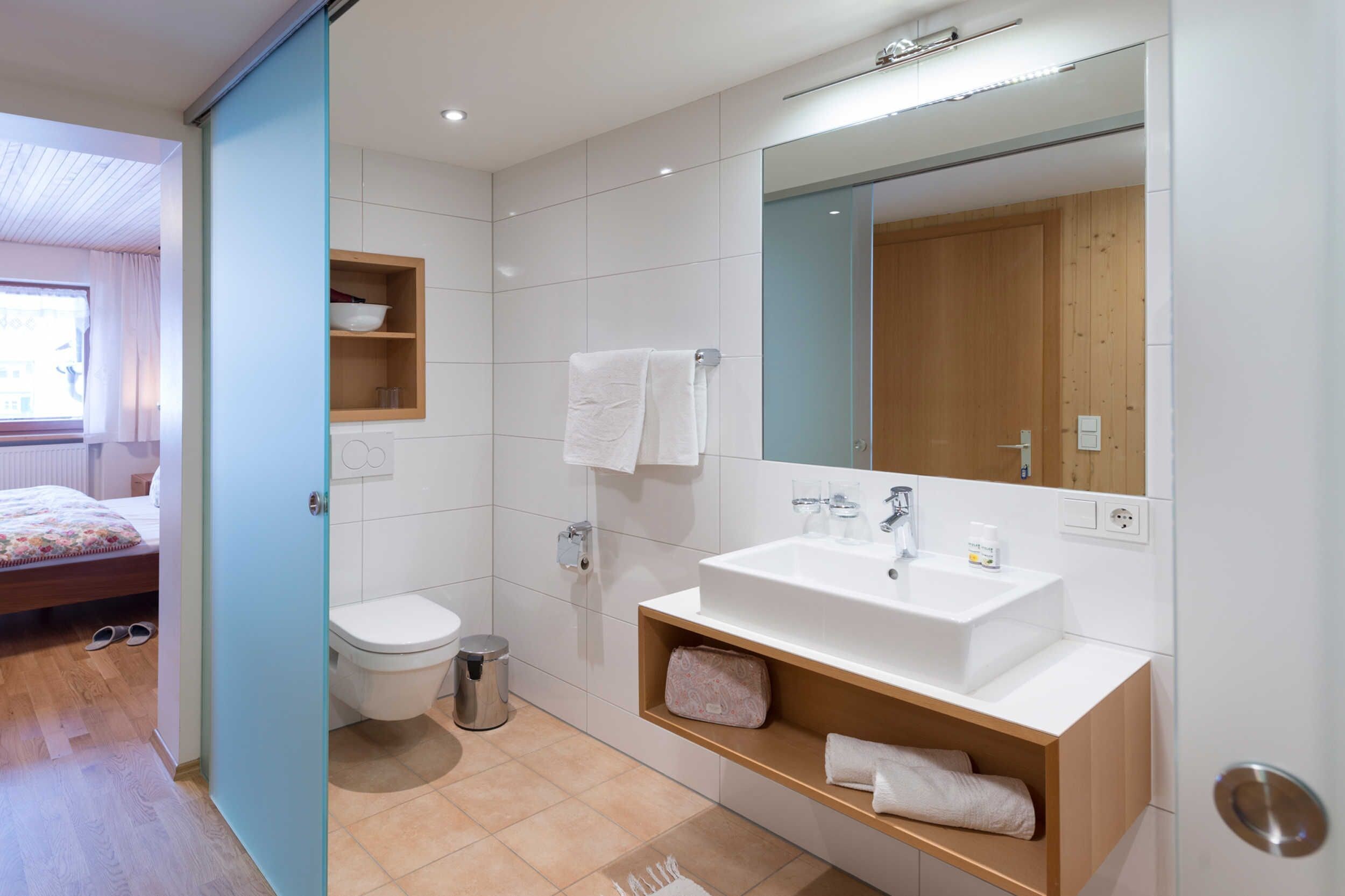 Ein modernes Badezimmer mit einem weißen Waschbecken, Spiegel und WC. Ein Holzregal beherbergt Handtücher und einen Mülleimer.