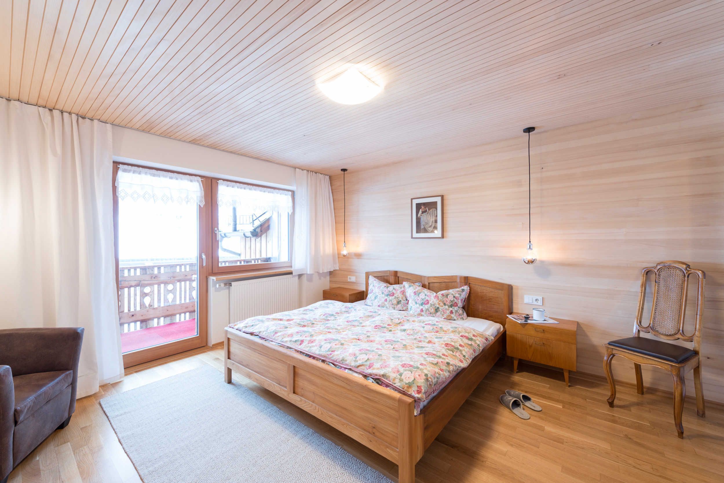 Ein Schlafzimmer mit einem hölzernen Bett, einem Nachttisch, einem Sessel, einem Fenster mit Vorhängen und Blick auf einen Balkon.