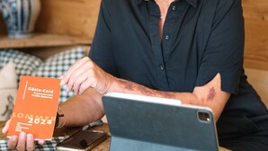Uma pessoa sentada a uma mesa de madeira com um laptop, segurando um cartão com a inscrição 'Gästecard Sommer 2024'.