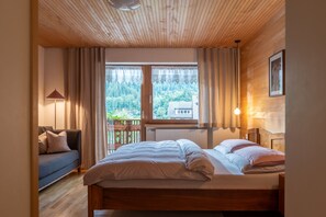 Un dormitorio con techo de madera, una cama con ropa de cama blanca, una mesita de noche, una ventana con cortinas y vistas a árboles y edificios.