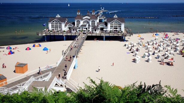 A pier leading to a white building with multiple windows, a sandy beach, and umbrellas. - Vacation home 04 and terrace - Holiday apartments with terrace in Sellin for (Sellin)