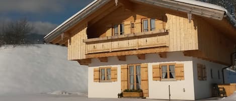 A wooden chalet with a balcony, large windows, and a snow-covered ground.