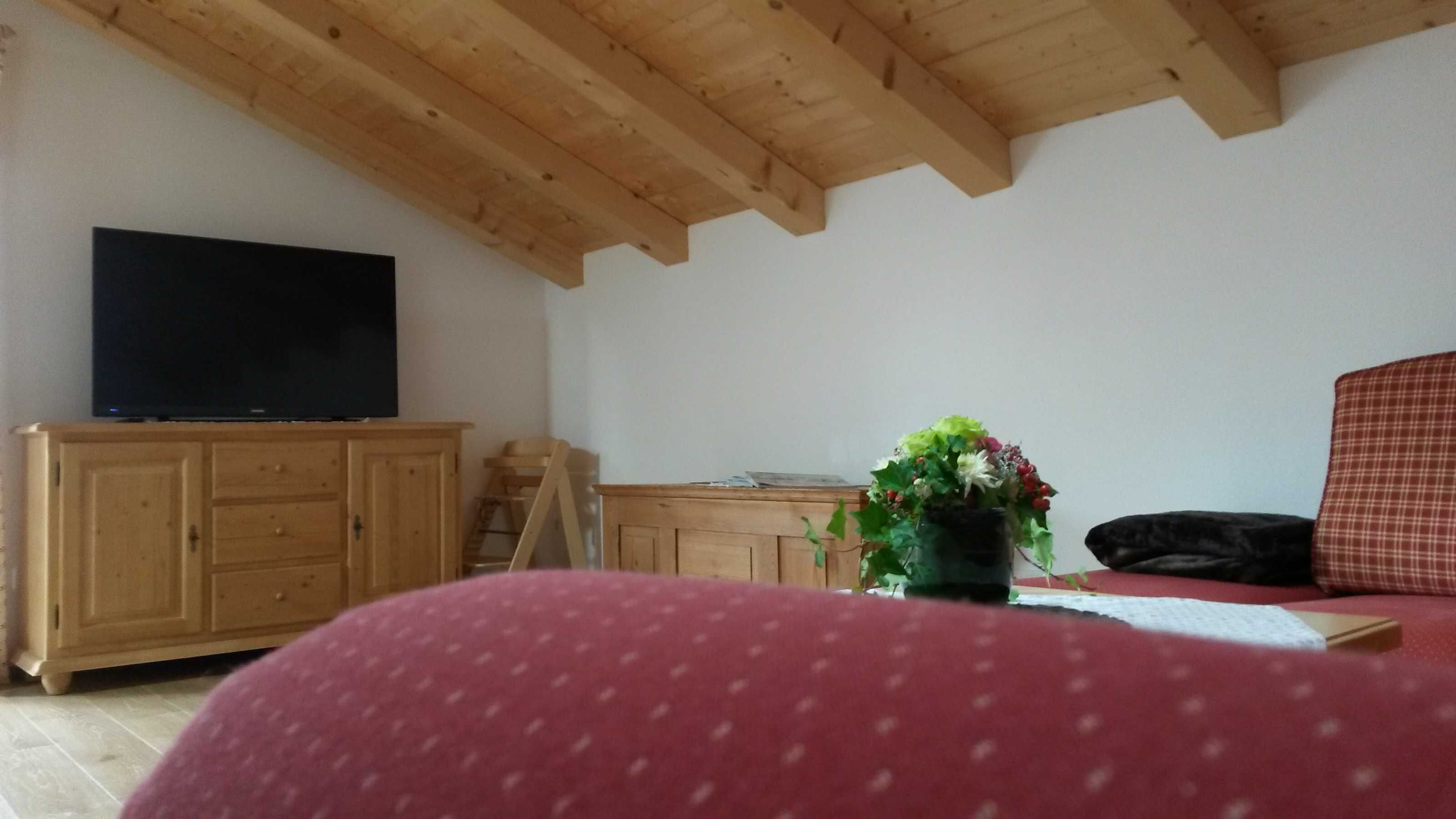 Una habitación con techo de madera, un televisor sobre un mueble, un sofá rojo y una pequeña mesa con un florero.