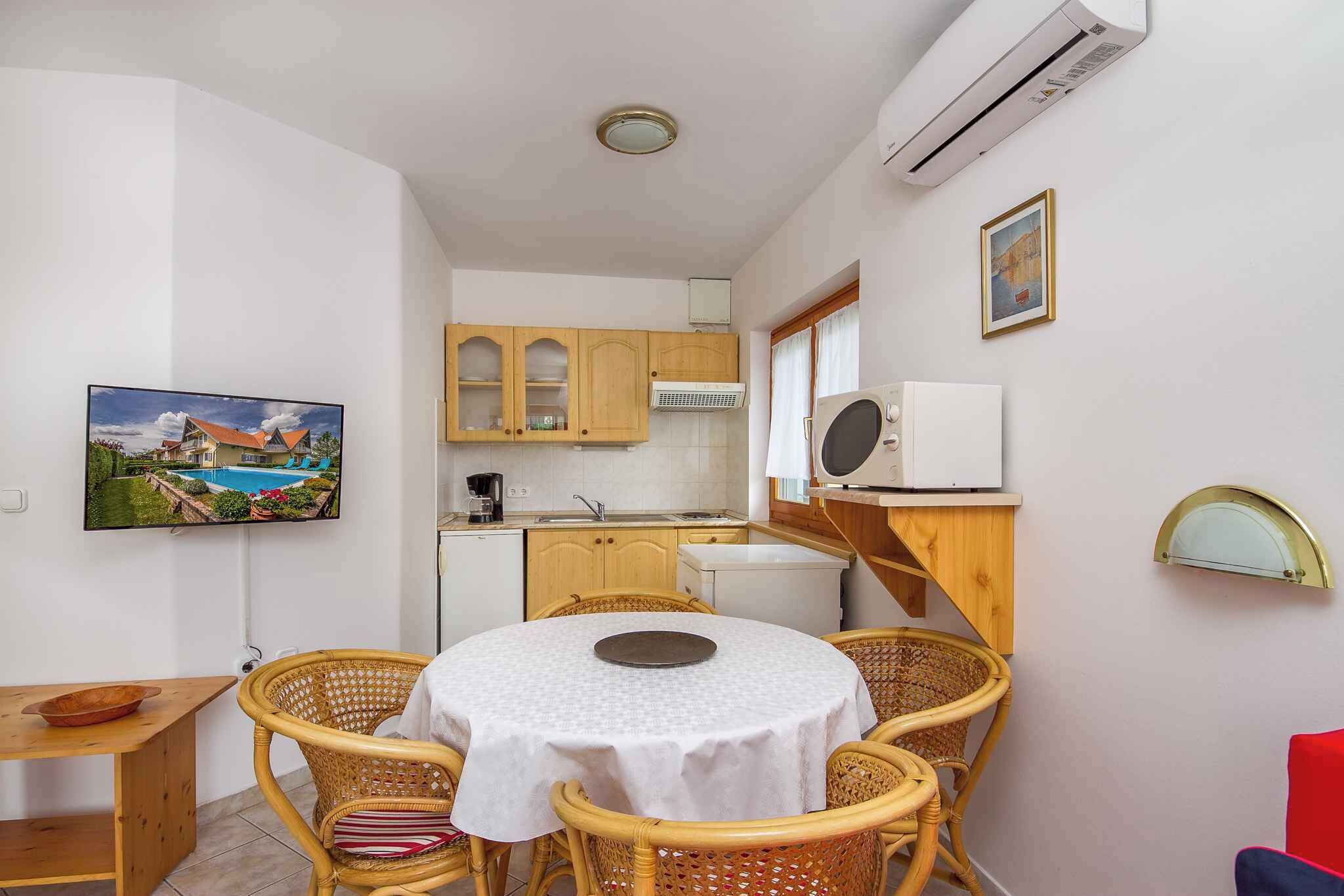 Cocina compacta con armarios de madera, comedor con sillas de mimbre y televisor montado en la pared.