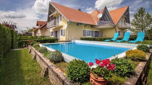 Una casa de dos pisos con piscina, sillas de descanso azules y un jardín bien cuidado.