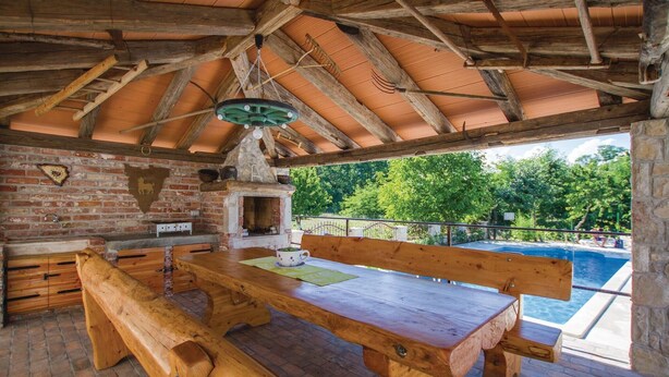 Un comedor al aire libre rústico con pared de ladrillo, bancos de madera y vista a una piscina.