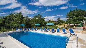 Una piscina con agua azul, rodeada de sillas de descanso y sombrillas, con árboles y un cielo despejado al fondo.