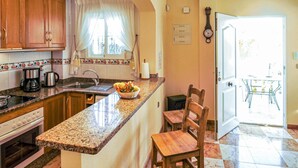 Cuina amb encimera de granit, armaris de fusta, un rentapeus i una cuina. Hi ha una finestra amb cortines, un rellotge a la paret i una porta que dóna a una zona de menjador.