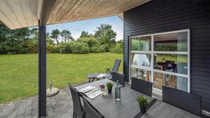 Ein modernes Haus mit überdachter Terrasse, Outdoor-Möbeln, einer Schaukel und Blick auf die umliegende Natur.