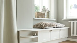 A white built-in bench with storage drawers, a wicker basket, and a decorative item on a window sill. - Apartment 01 - "Villa Minerva" Apartment 01 (Kühlungsborn)