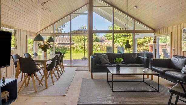 A spacious living area with a high ceiling, large windows, a dining area with chairs, a sofa, and a coffee table. - Nice home in Væggerløse with sauna (Væggerløse)