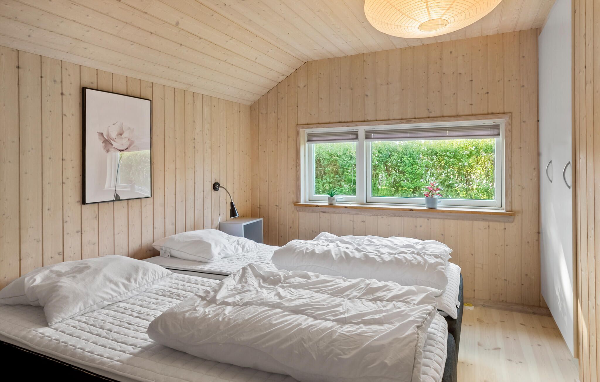 A bedroom with a wooden ceiling and walls, a bed with white bedding, a bedside table with a lamp, and a window with a view of greenery.