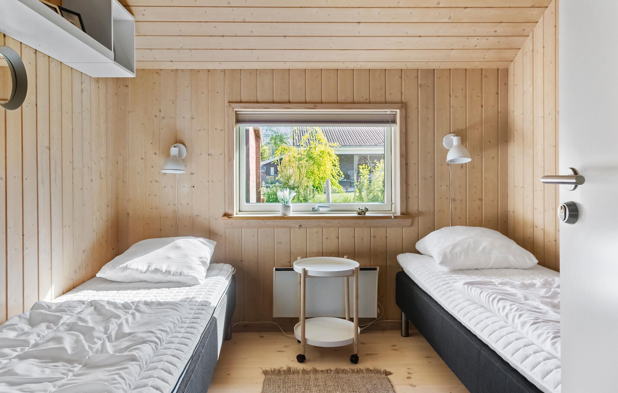 A small, cozy bedroom with two single beds, a small table, and a window with a view of greenery outside.