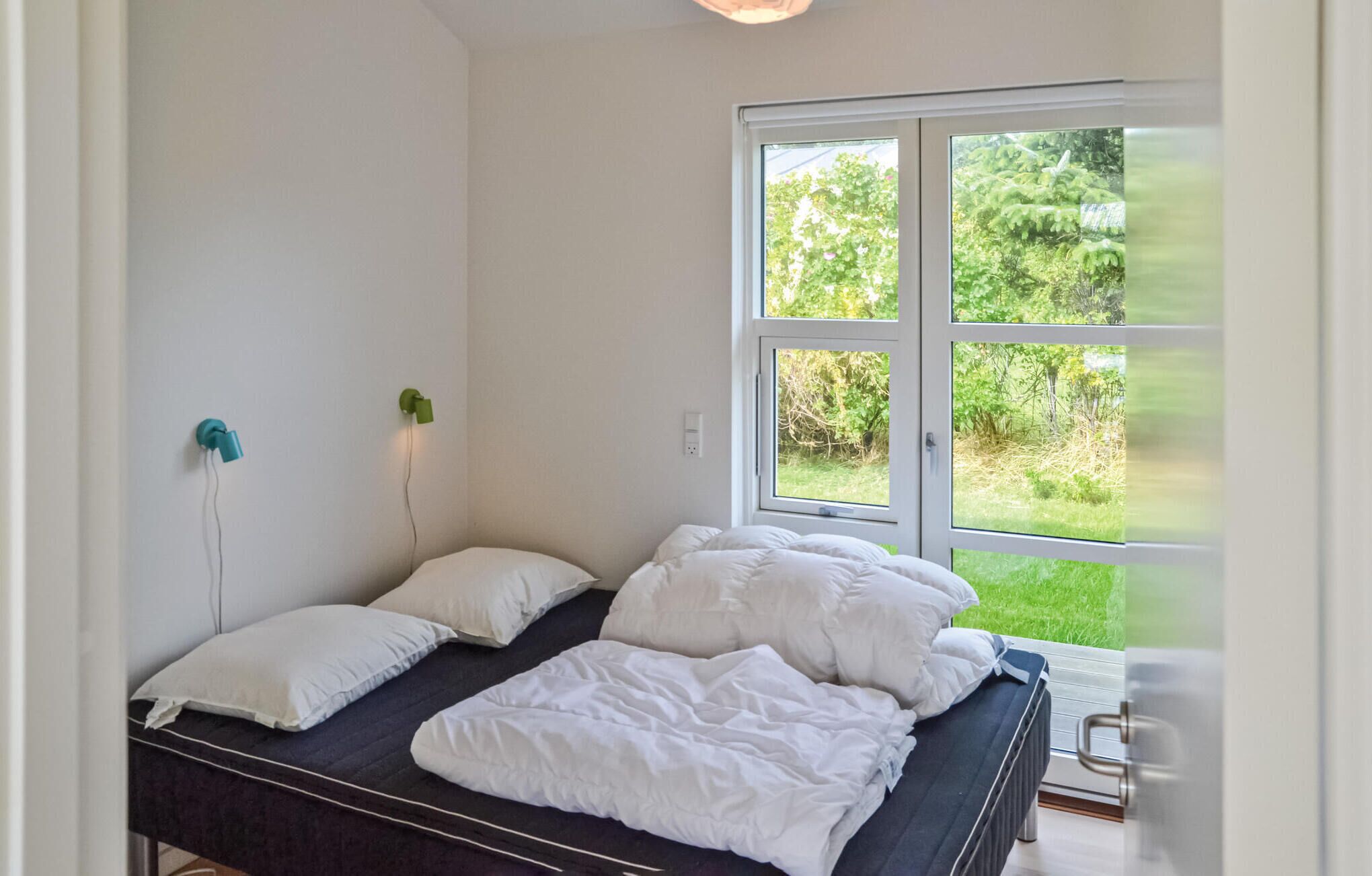 Een slaapkamer met een bed, witte kussens, een nachtkastje, een groene wandlamp en een groot raam met uitzicht op groen.