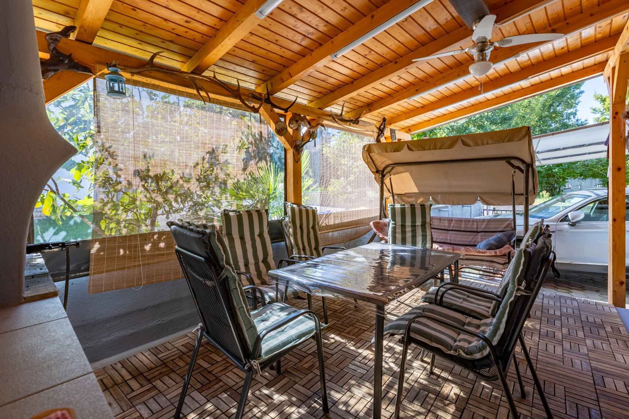 Een terras buiten met een houten dak, een tafel en strepenstoelen.