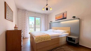Een slaapkamer met een bed, nachtkastje, bureau, stoel, raam met gordijnen, schilderij en een kroonluchter.