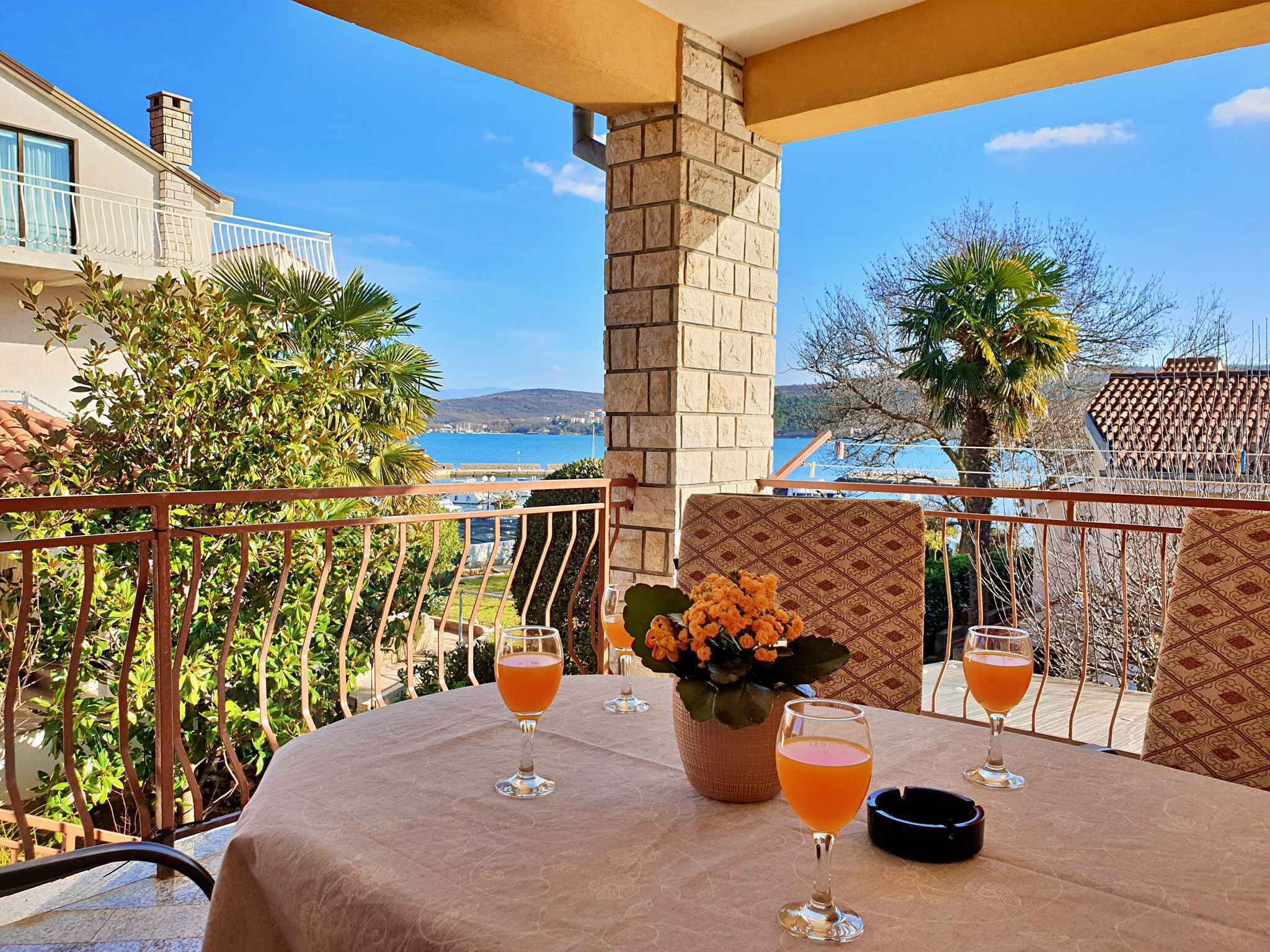 Een balkon met een tafel gedekt voor twee personen, met een vaas met bloemen en twee glazen oranje drank.