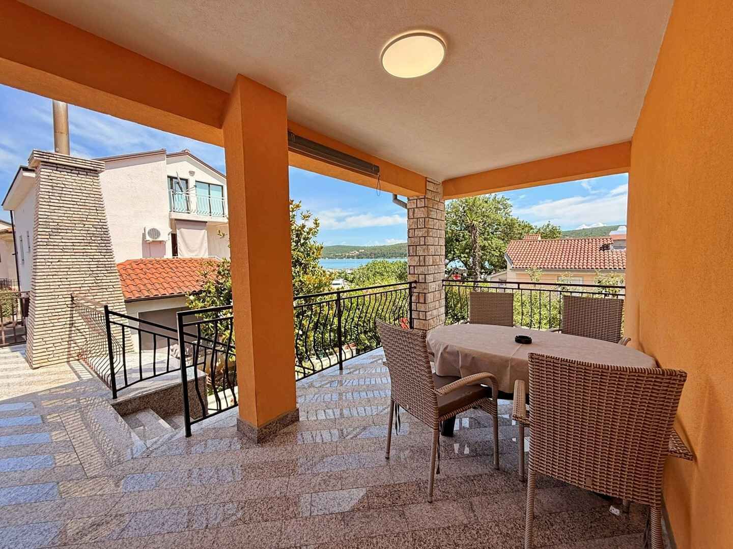 En balkon med bord og stole, havudsigt og en bygning med et tagtækket tag i baggrunden.