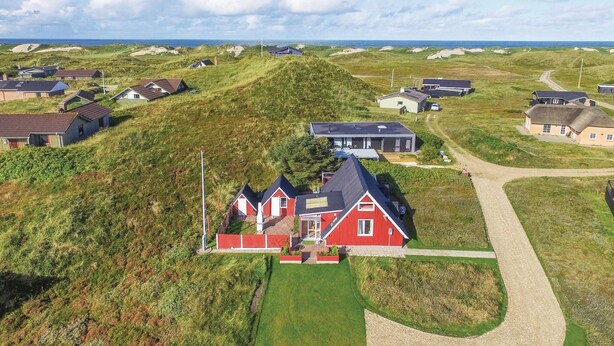 A coastal house with a red facade, surrounded by grassy dunes and a gravel driveway.