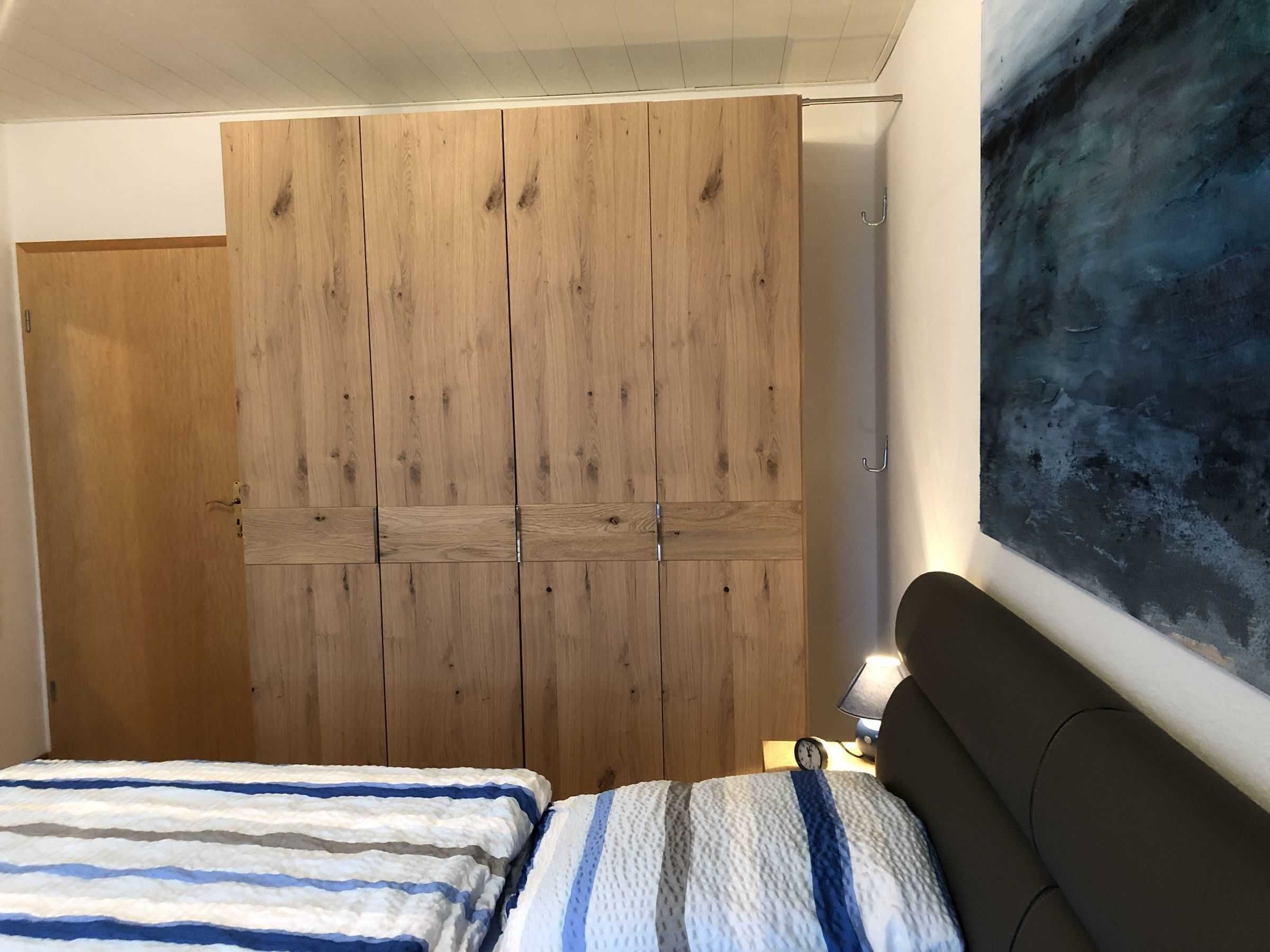 A bedroom with a wooden wardrobe, a bed with striped bedding, and a wall-mounted lamp.