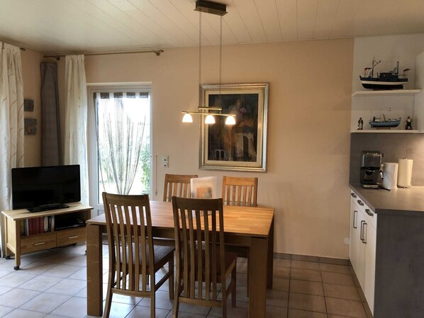 Un coin repas avec une table et des chaises en bois, un téléviseur posé sur un meuble, un tableau accroché au mur, et un espace cuisine équipé d’une machine à café et d’étagères.