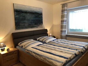 A bedroom with a bed, a nightstand, a lamp, a painting, and a window with curtains.