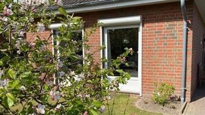 A brick building with a white door and a small garden area with flowering plants. - Vacation cottage Seestern, Germany (Friedrichskoog)