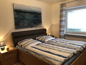 A bedroom with a bed, a nightstand, a lamp, a painting, and a window with curtains. - Vacation cottage Seestern, Germany (Friedrichskoog)
