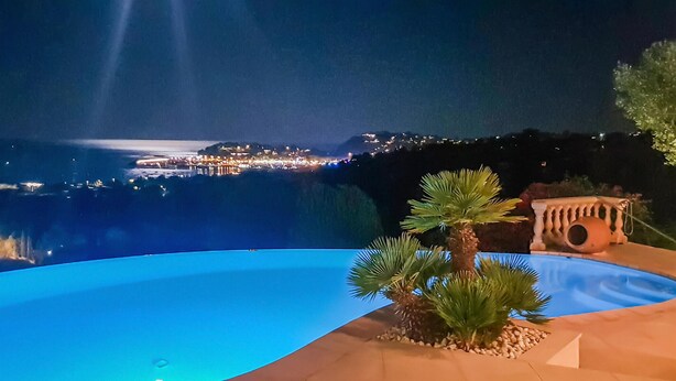 Vista nocturna de una zona costera con piscina, palmeras y un paisaje urbano distante.