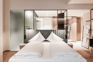 A modern hotel room with a large bed, a mirror, a wooden wardrobe, and a bathroom visible through a glass partition.