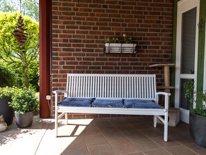 Un banc blanc avec des coussins bleus, placé sur une terrasse carrelée, adossé à un mur de briques, et agrémenté de plantes en pot.