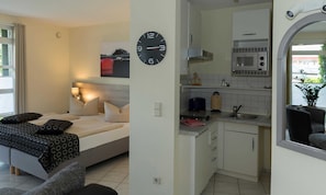 Une chambre à coucher avec un lit, des oreillers, une table de chevet et une horloge murale. La pièce adjacente est une cuisine équipée d’un four à micro-ondes, d’un évier, de placards et d’une fenêtre.