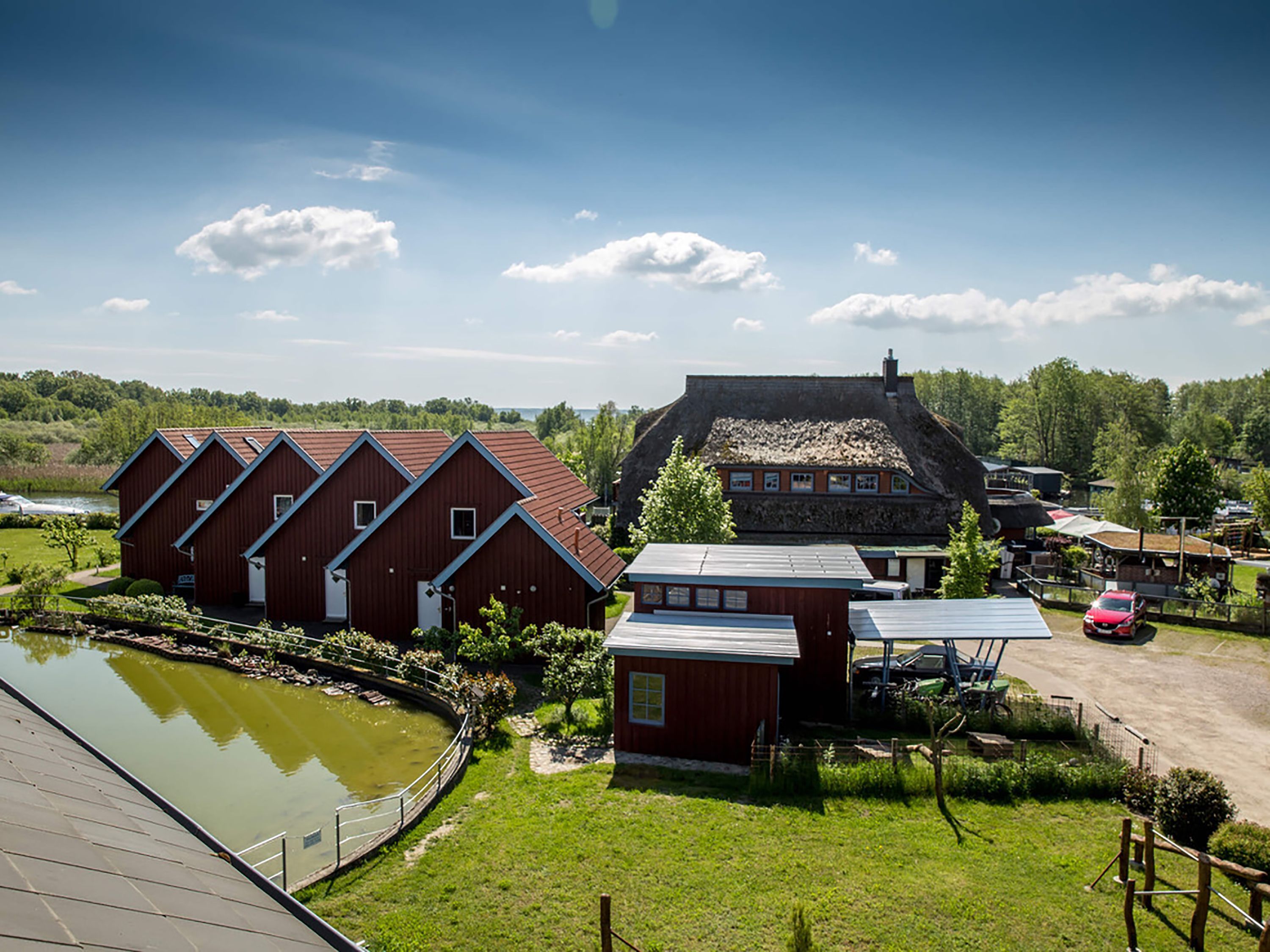 Un paysage rural avec une rangée de bâtiments en bois peints en rouge, un étang et une maison à toit de chaume.