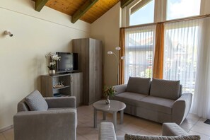 Un salon comprenant un canapé, un fauteuil, un téléviseur, un meuble et une petite table sur laquelle est posé un vase.