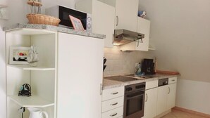 A compact kitchen with white cabinets, a built-in oven, and a microwave. There are shelves with kitchenware and a coffee machine on the countertop. - (177) 2-room apartment Ostseeallee - apartment house dune castle (Kühlungsborn)