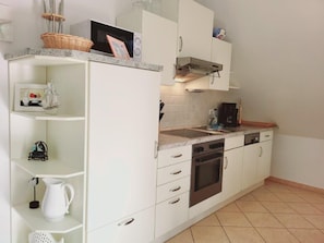 A compact kitchen with white cabinets, a built-in oven, and a microwave. There are shelves with kitchenware and a coffee machine on the countertop. - (177) 2-room apartment Ostseeallee - apartment house dune castle (Kühlungsborn)
