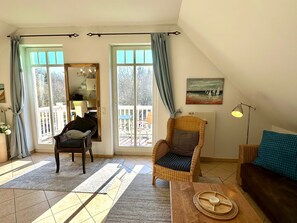 A cozy living room with a sofa, a chair, and a dining table. There is a large window with curtains, a painting on the wall, and a view of the outdoors. - (177) 2-room apartment Ostseeallee - apartment house dune castle (Kühlungsborn)
