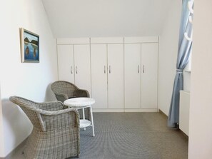 A room with a high ceiling, a white wardrobe, two wicker chairs, a small round table, and a painting on the wall.
