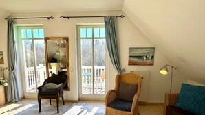 A cozy living room with a sofa, a chair, and a dining table. There is a large window with curtains, a painting on the wall, and a view of the outdoors. - (177) 2-room apartment Ostseeallee - apartment house dune castle (Kühlungsborn)
