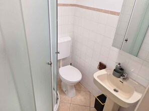 A compact bathroom with a toilet, sink, and mirror, featuring a tiled interior and a door leading to the adjacent room.