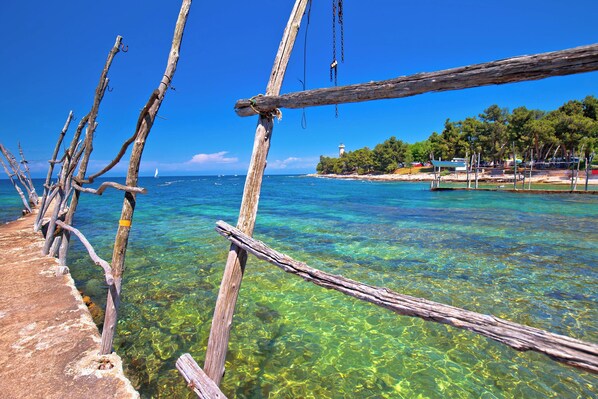 A wooden pier extending into clear blue waters with a lighthouse and boats in the distance.