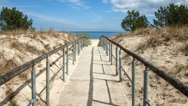 Ein hölzerner Steg führt zu einem Strand, beidseitig mit Metallgeländern.