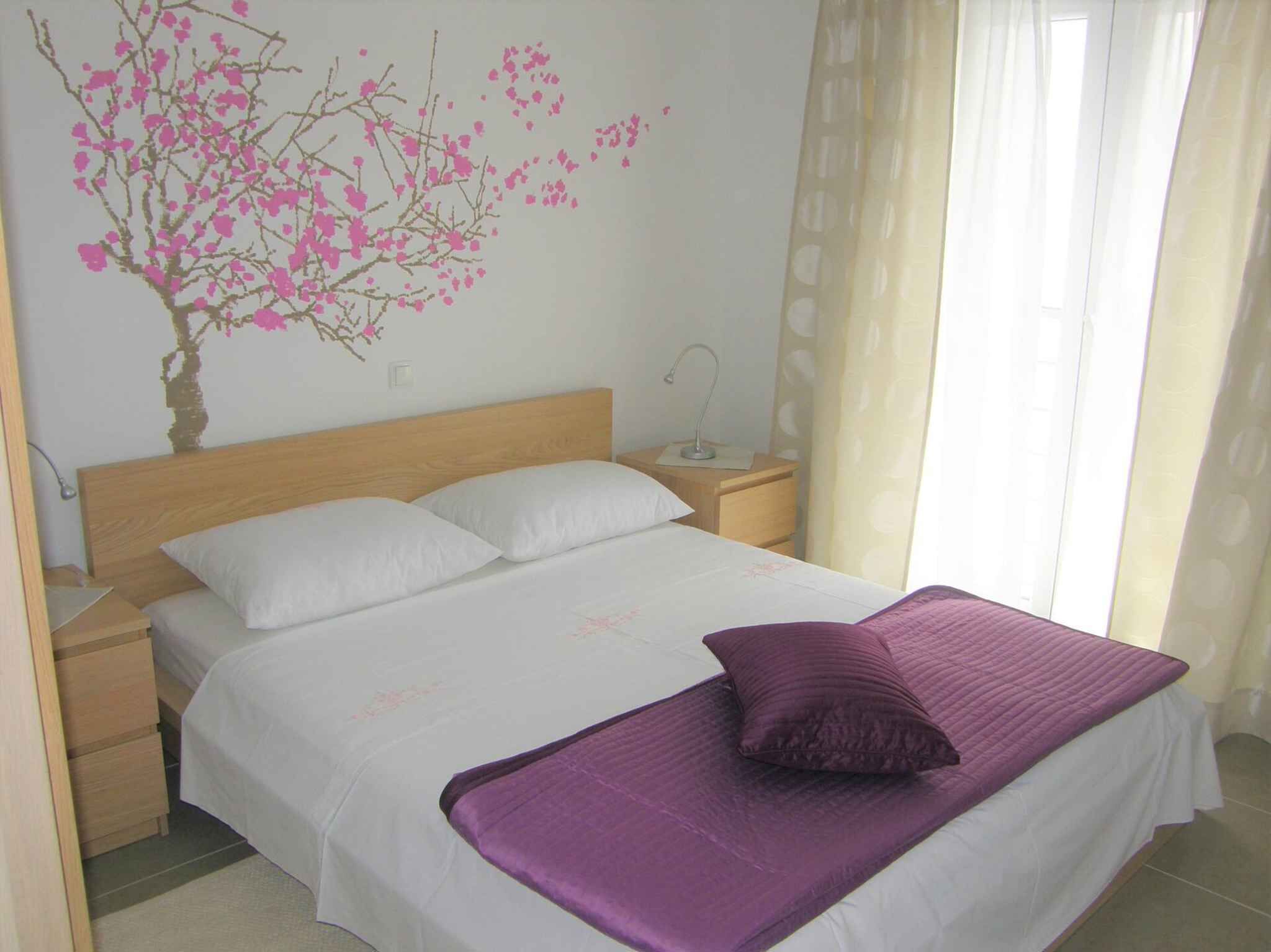 A bedroom with a bed, white bedding, a purple throw, a nightstand, a lamp, and a wall decoration of pink flowers.