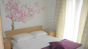 Ein Schlafzimmer mit einem Bett, weißer Bettwäsche, einer violetten Tagesdecke, einem Nachttisch, einer Lampe und einer Wanddekoration mit rosafarbenen Blumen.