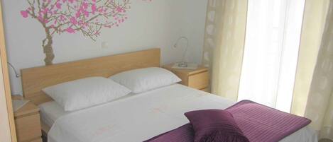 A bedroom with a bed, white bedding, a purple throw, a nightstand, a lamp, and a wall decoration of pink flowers.