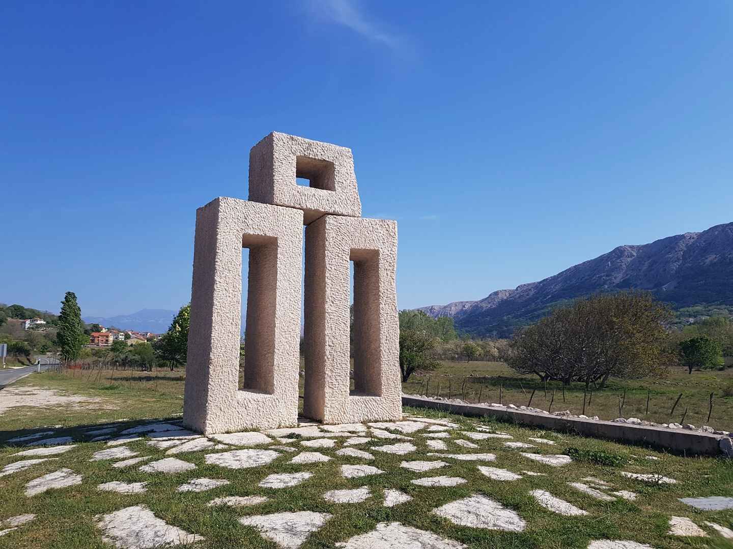 A stone monument with a square top and rectangular sides, set on a patterned stone pavement, surrounded by grass and trees, with mountains in the background.