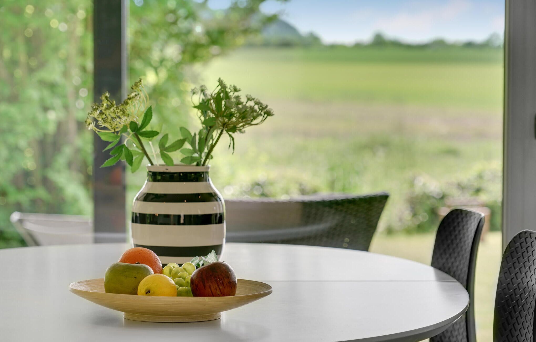 Une table ronde à la surface blanche, un saladier en bois rempli de fruits et un vase rayé avec des fleurs.